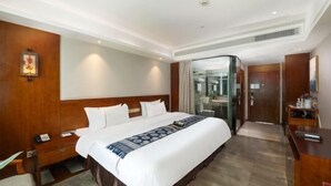 A hotel room with a large bed, a bedside table with a phone, a mirror, and a glass-enclosed bathroom. - Harmonious Qianjin Hotel (Kaili)