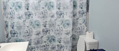 Un baño con cortina de ducha estampada con motivos florales, un lavamanos blanco, un inodoro y un carro con toallas.