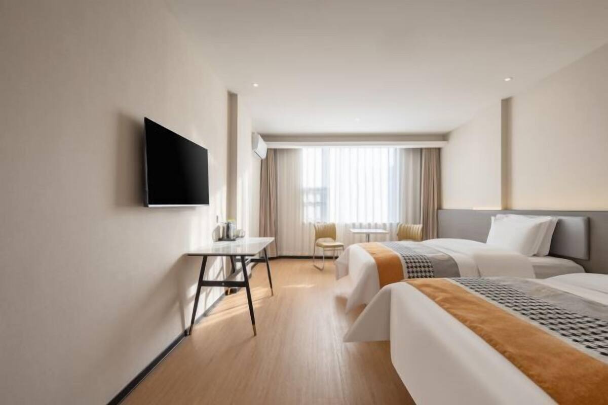 A hotel room with a bed, a desk, a chair, a TV mounted on the wall, and a window with curtains.