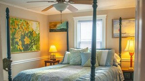 A four-poster bed with a ceiling fan, a nightstand with a lamp, a painting on the wall, and a window with blinds. - Cozy Rehoboth Beach 2BR Retreat - Just 1.5 Blocks to the Beach! (Rehoboth Beach)
