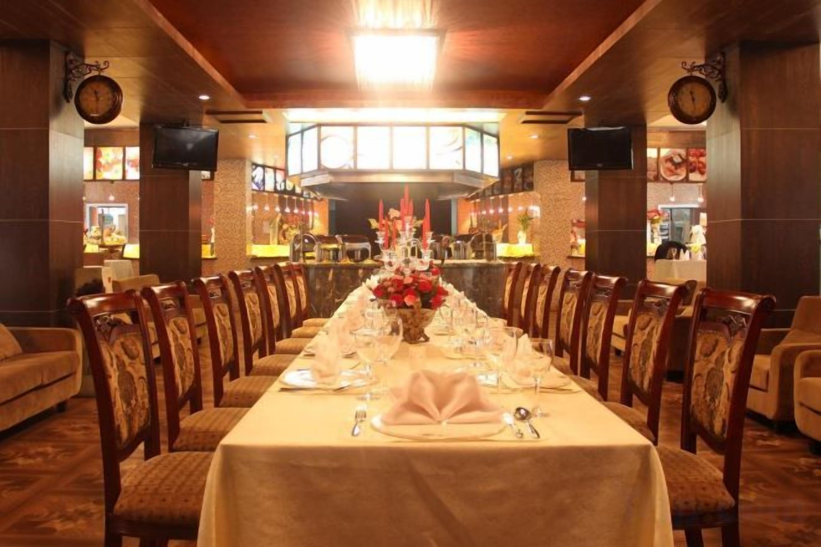 Una larga mesa de comedor dispuesta para una comida formal en el comedor de un hotel, con múltiples sillas, un reloj en la pared y una pantalla de televisión.