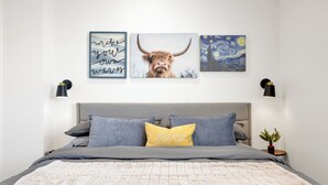 Une chambre à coucher avec un lit, des oreillers, une table de chevet ornée d’une plante et des œuvres d’art murales, dont une représentation d’une vache et une reproduction d’un tableau célèbre.