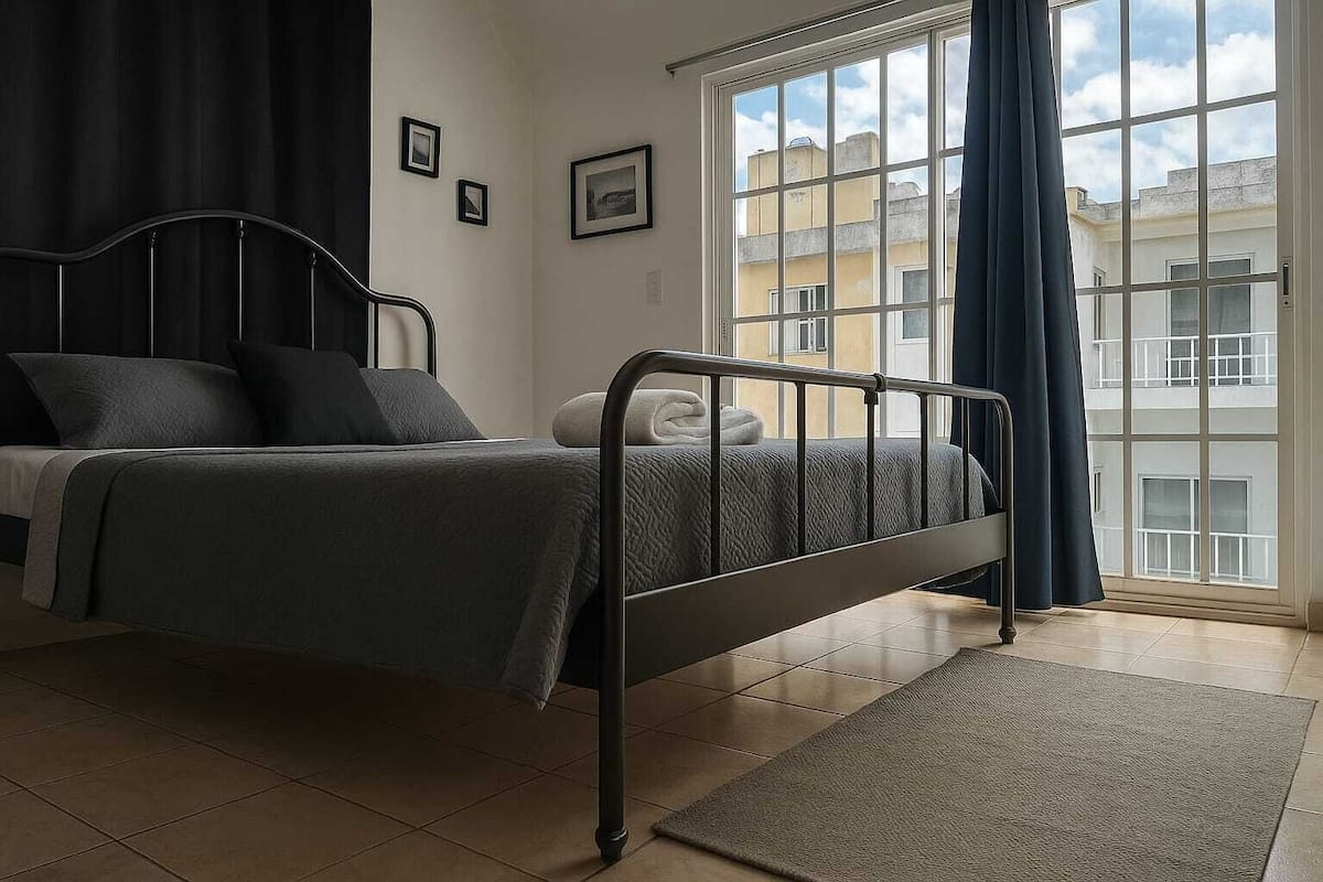 A bedroom with a bed, a window with curtains, and a view of buildings outside.