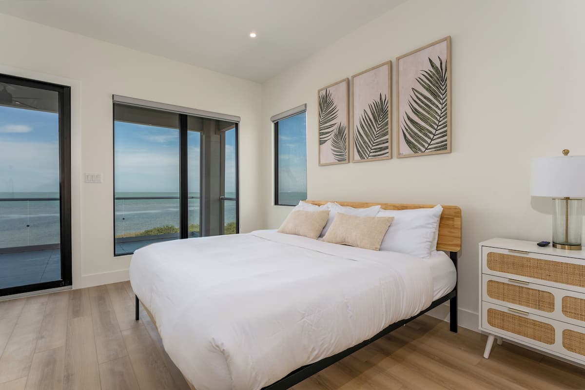 A bedroom with a large bed, a nightstand, and a view of the ocean through large windows.