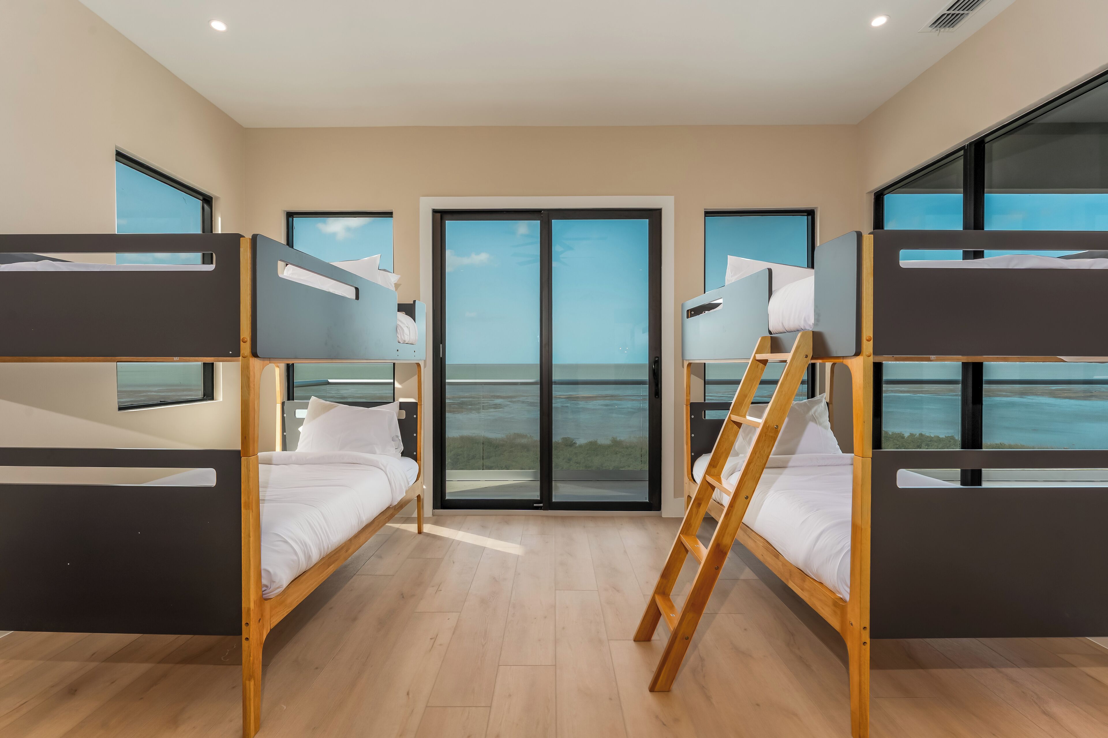 A room with bunk beds, a wooden ladder, and large windows offering a view of the sea.