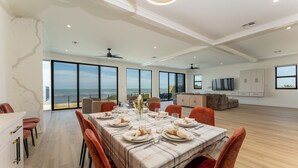 Uma ampla área de refeições com uma grande mesa posta para seis, uma cozinha ao fundo e vista para o oceano através de grandes janelas.