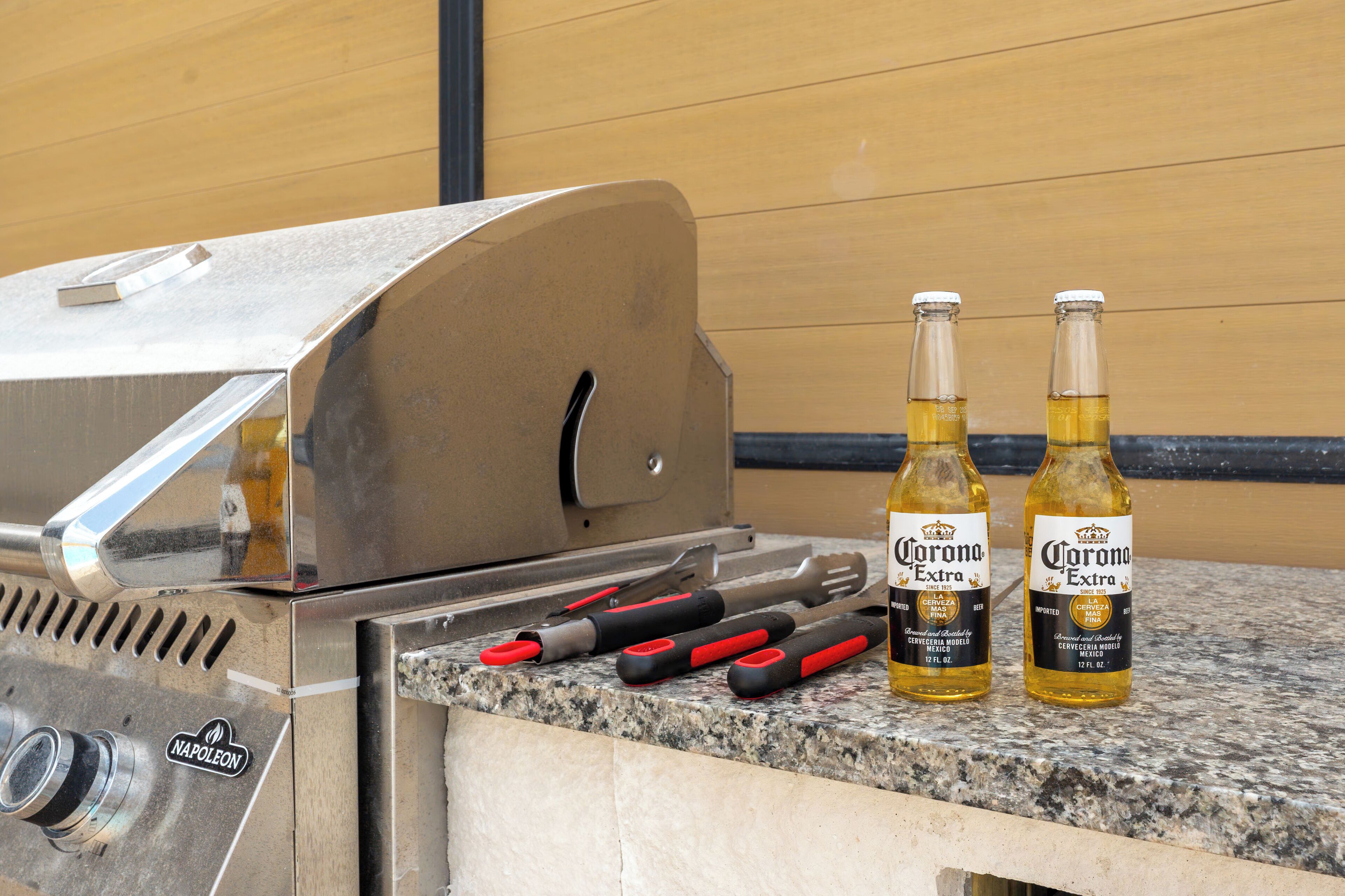 A stainless steel grill with two bottles of Corona Extra beer and barbecue tools on a granite countertop.