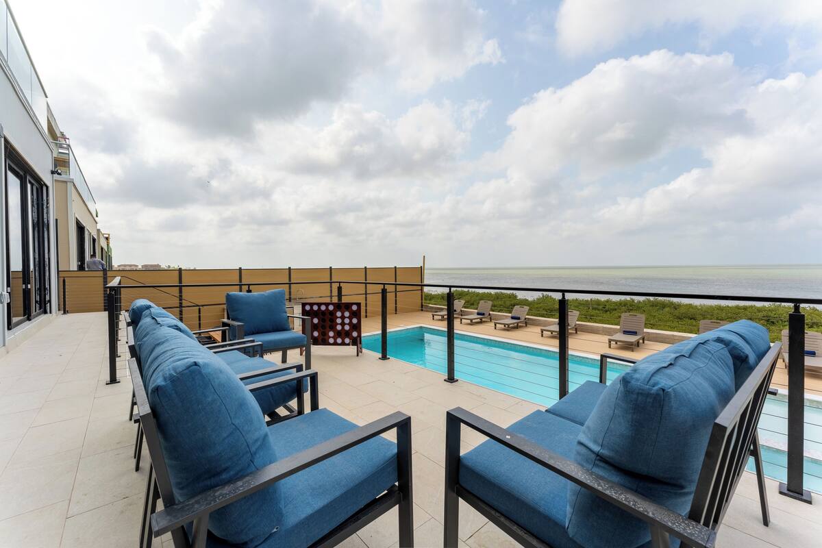 A rooftop pool area with blue cushioned chairs and a view of the sea.