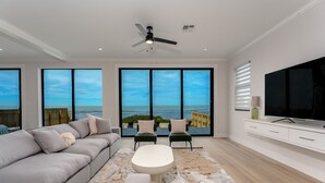 Uma sala de estar moderna com um sofá grande, uma mesa de centro e uma TV sobre um armário branco. Há duas poltronas e uma janela ampla com vista para o oceano.