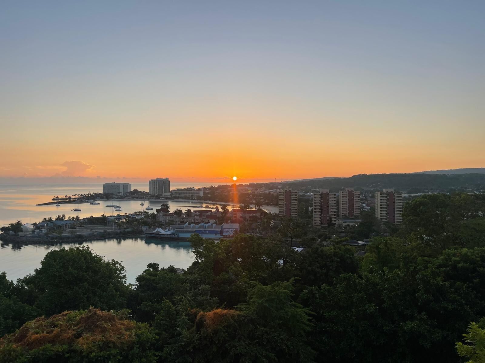 Une vue d’une ville côtière au coucher du soleil, avec des bâtiments, un port de plaisance et une zone boisée.