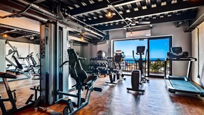 A gym with various exercise equipment, a large window with a view of the sea, and a ceiling with exposed pipes.