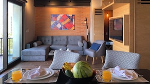 A modern living room with a gray sofa, patterned chairs, a dining table with plates and a watermelon, and a large abstract painting on the brick wall.