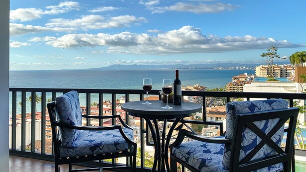 A balcony with a table and two chairs, overlooking a coastal view with buildings and a clear sky.