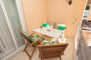 Ein Balkon mit einem weißen Tisch, zwei Stühlen mit grünen Kissen mit Blattmuster, einem kleinen Topfpflanzen und einer weißen Wand.