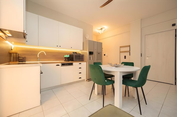 Une cuisine moderne avec des meubles blancs, un micro-ondes encastré et un coin repas comprenant une table blanche et des chaises vertes.