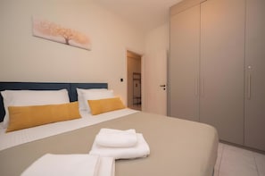 Un lit bien fait, avec des oreillers blancs et moutarde, un tableau représentant un arbre accroché au mur et une armoire fermée.