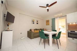 Una sala de estar moderna con un sofá, una mesa de comedor con sillas, un ventilador de techo, un televisor montado en la pared y una zona de cocina con horno.