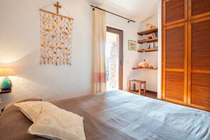 A bedroom with a bed, a wall-mounted shelf with books, a wooden wardrobe, and a window with curtains.