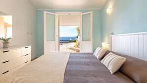 A bedroom with a bed, a dresser, a nightstand, and a view of the sea through an open door.