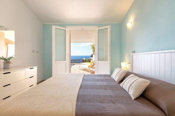 A bedroom with a bed, a dresser, a nightstand, and a view of the sea through an open door.