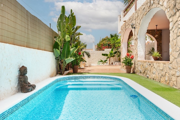 Une piscine dont le bord est recouvert de carreaux, entourée d’un mur de pierre, de plantes en pot et d’une statue de Bouddha.