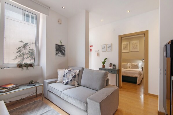 Un salon moderne avec un canapé, une petite table, une fenêtre ornée d’une plante et une porte donnant sur une chambre.