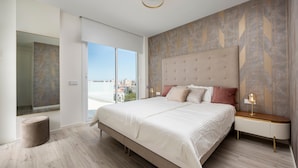 Un dormitorio moderno con una cama grande, una mesita de noche con lámpara, un taburete redondo y vistas de la ciudad a través de un ventanal amplio.