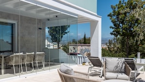Un espacio exterior moderno con mobiliario de mimbre y una pared de vidrio con vistas a un paisaje urbano.