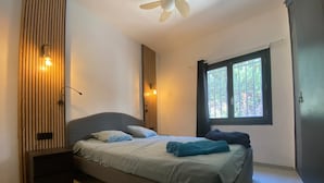 A bedroom with a bed, a ceiling fan, a window with a view of trees, and a wall-mounted light fixture.