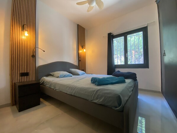 A bedroom with a bed, a ceiling fan, a window with a view of trees, and a wall-mounted light fixture.