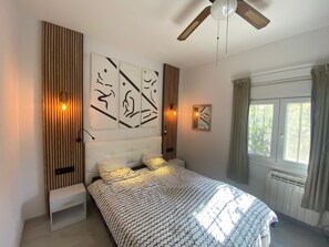 Une chambre à coucher avec un lit, un ventilateur de plafond, des œuvres d’art accrochées au mur et une fenêtre avec des rideaux.