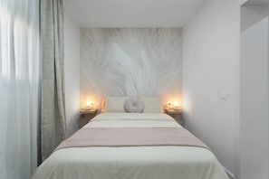 Un dormitorio con una cama, dos mesitas de noche, un buró, una lámpara de pared y un ventanal con cortinas.