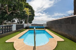 Una piscina con barandilla de metal, rodeada de césped artificial y un muro de piedra.