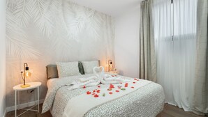 Een keurig opgemaakt bed met een beddengoed met patroon, twee nachtkastjes met lampen, en een groot raam met dunne gordijnen.