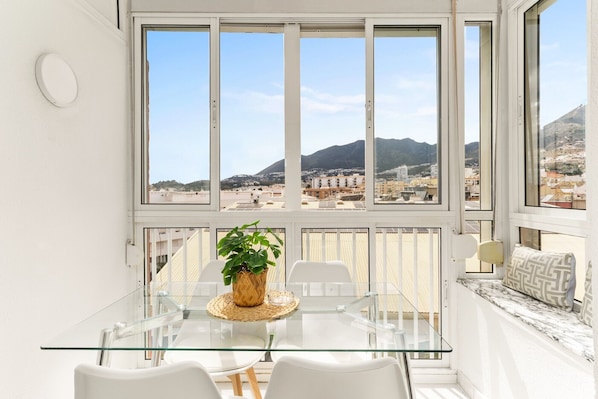 A modern room with a glass table, white chairs, a potted plant, and a view of a cityscape with mountains.