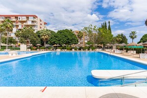 A swimming pool with lounge chairs and umbrellas, surrounded by greenery and a multi-story building.