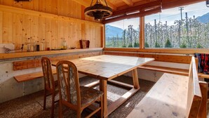Un interno in legno con un tavolo e panche, un lampadario e una finestra che si affaccia su alberi e montagne.