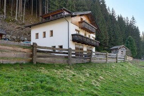 Un chalet de dos pisos con balcón de madera, rodeado por una cerca de madera y con un bosque al fondo.