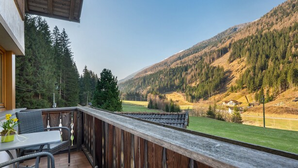 Ein Balkon mit Korbmöbeln, einem Hollerleger und Blick auf eine gebirgige Landschaft.