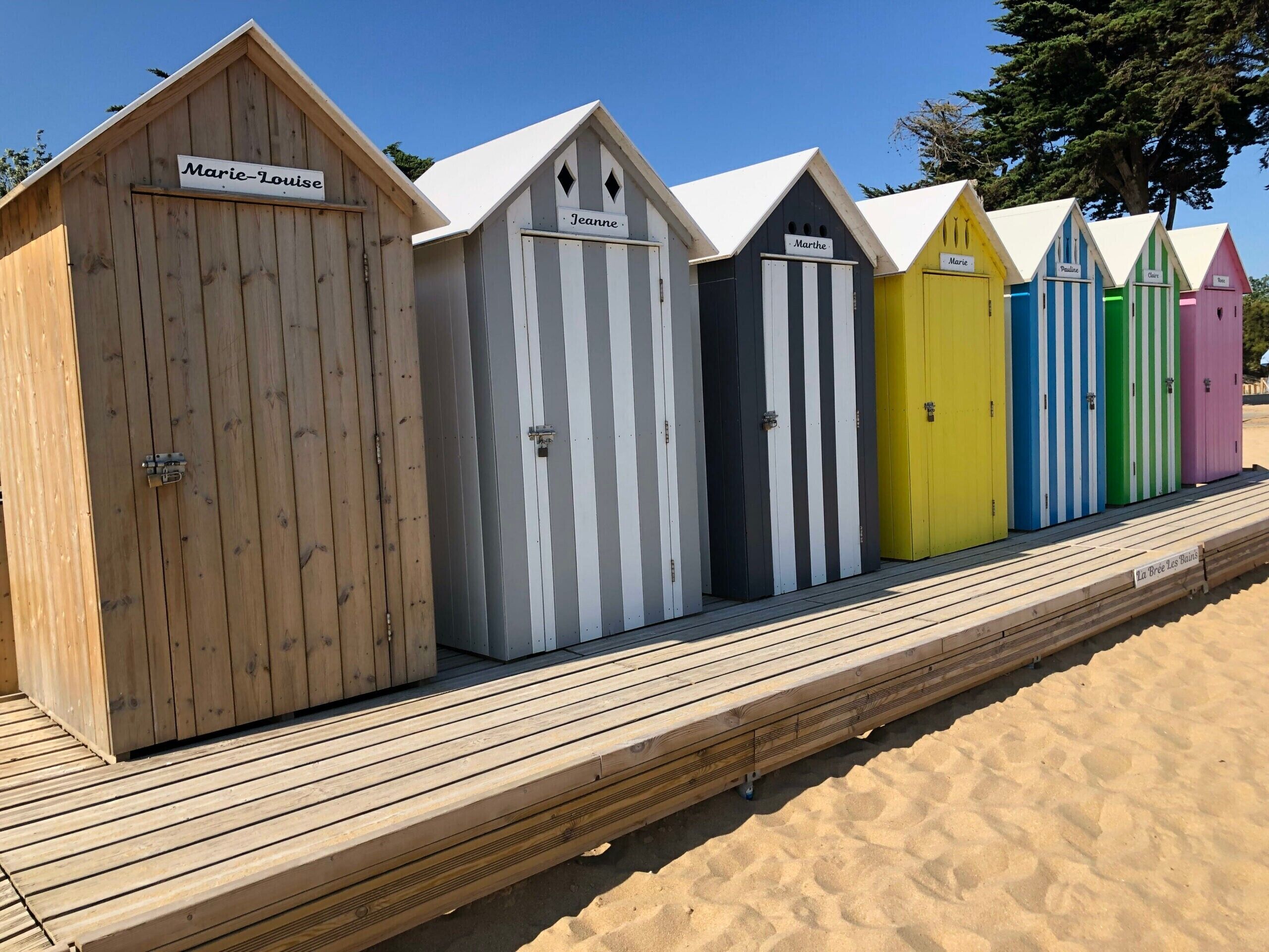 A row of beach huts with names on signs, wooden deck, and sandy beach.