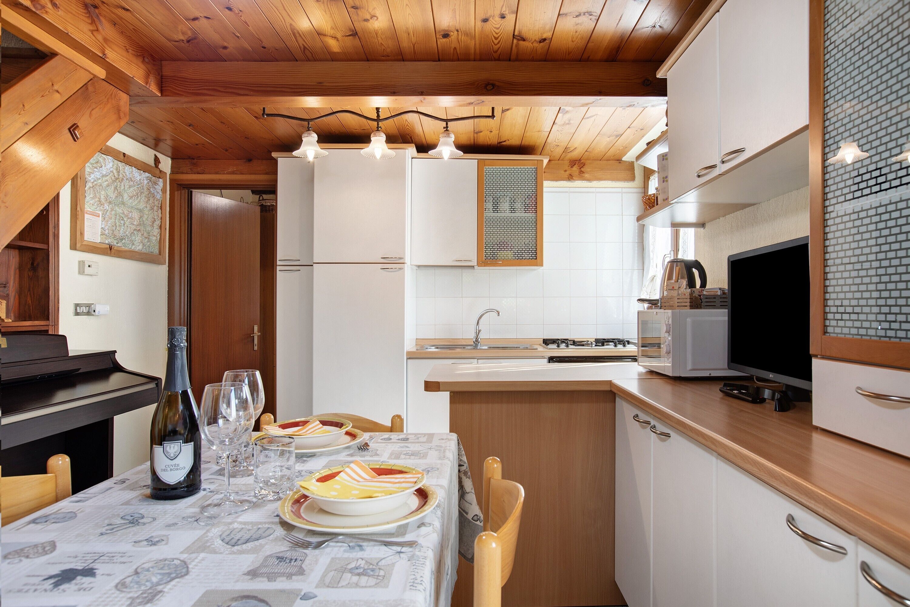 A kitchen with wooden cabinets, a dining table set for two, a wine bottle, and a television.