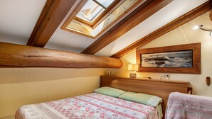 Un dormitorio con techo inclinado, vigas de madera, cama con ropa de cama estampada, un cuadro en la pared y una lámpara de noche.