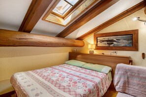 Une chambre avec un plafond en pente, des poutres en bois, un lit avec une literie à motifs, un tableau au mur et une lampe de chevet.