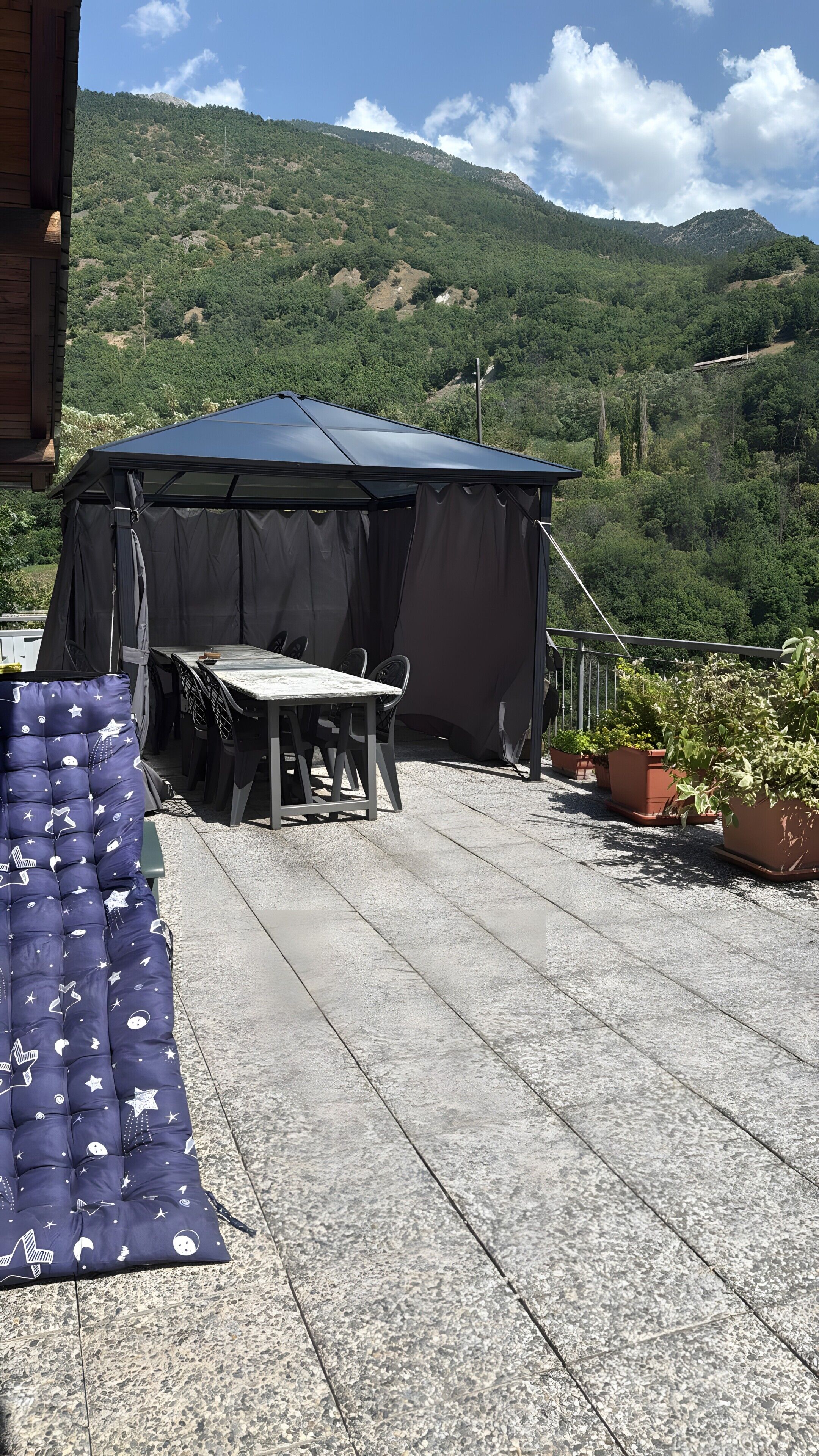 A covered outdoor seating area with a table and chairs, surrounded by potted plants and a view of a mountainous landscape.