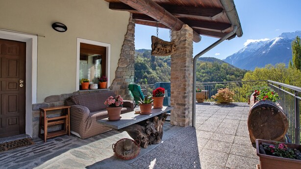 Un patio cubierto con pilares de piedra, un banco de madera, plantas en macetas y vistas a una montaña.