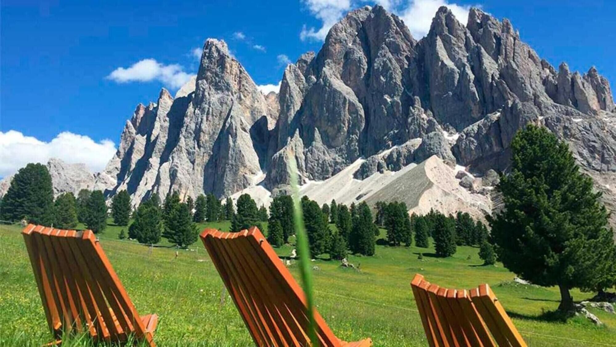 A mountain landscape with rocky peaks, a grassy field, and two wooden chairs.