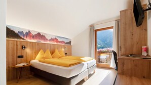 A bedroom with a bed, a nightstand, a wooden desk, a chair, and a large window with a mountain view. - Room 'Veltierhof 6' with Mountain View, Private Terrace and Wi-Fi (Funes)