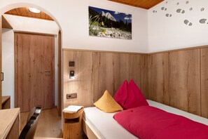 Een slaapkamer met een houten hoofdeinde, een bed met rode en gele kussens, een houten nachtkastje, een aan de muur hangend schilderij en een houten deur.