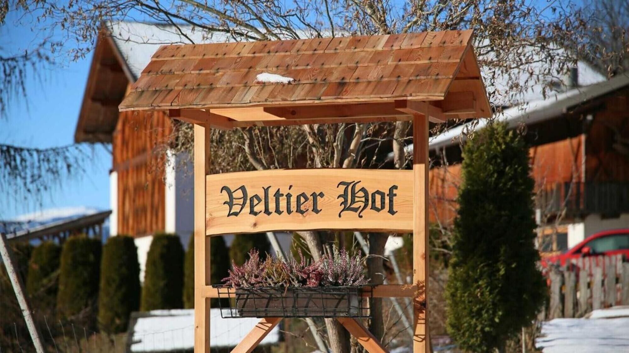 A wooden sign with the text 'Veltier Hof' and a planter with plants below it.