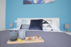 A neatly made bed with a blue wall and geometric pillows, a tray with mugs, plates, and cookies, and a small yellow flower.